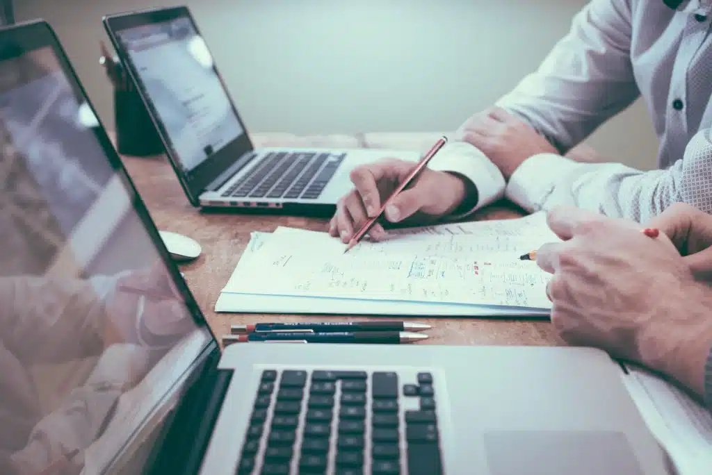 Mesa de trabalho com notebook e uma pessoa fazendo anotações em papel